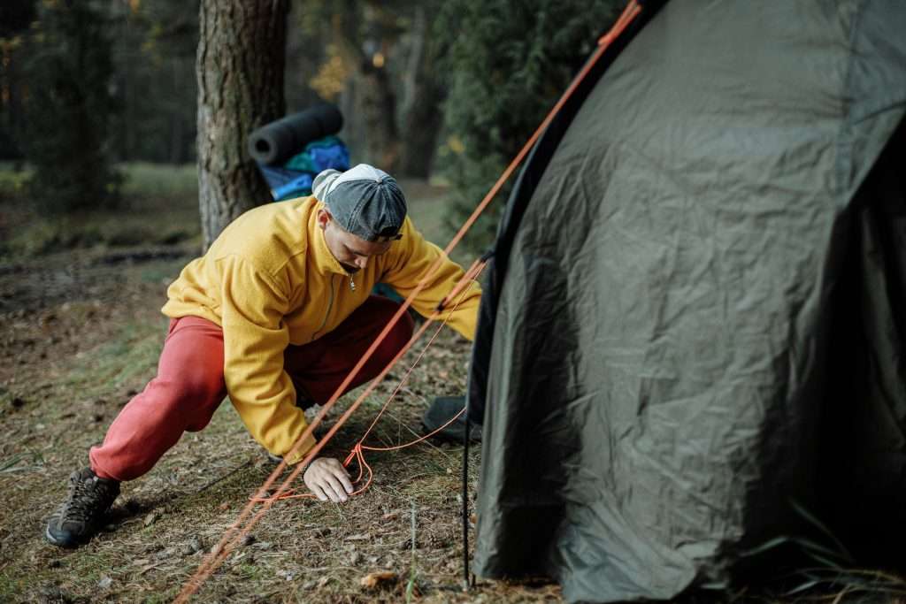 ragazzo con berretto e felpa gialla che sta montando una tenda canadese in un bosco