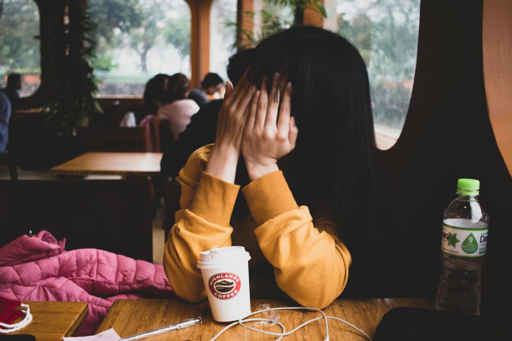 A woman in a yellow sweater covers her face at a cozy Hanoi coffee shop, surrounded by beverages and a warm ambiance. larp, timidezza nel larp